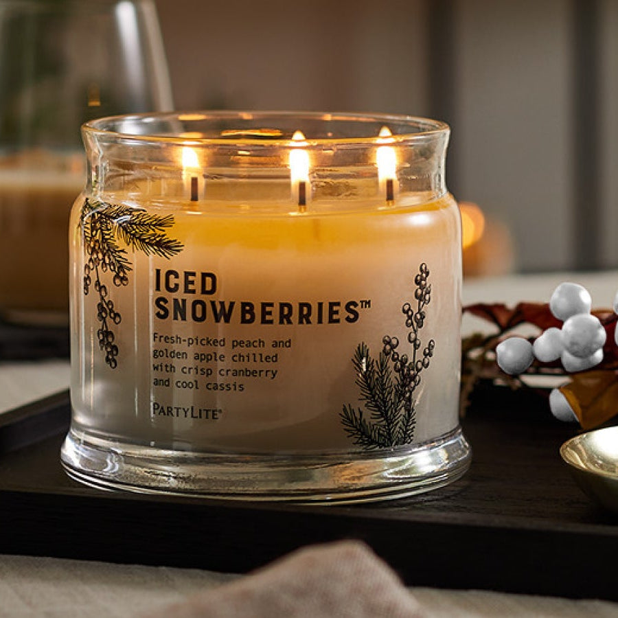 Lit off-white 3-wick candle on a tray next to red and white berries with two beverages in the background.