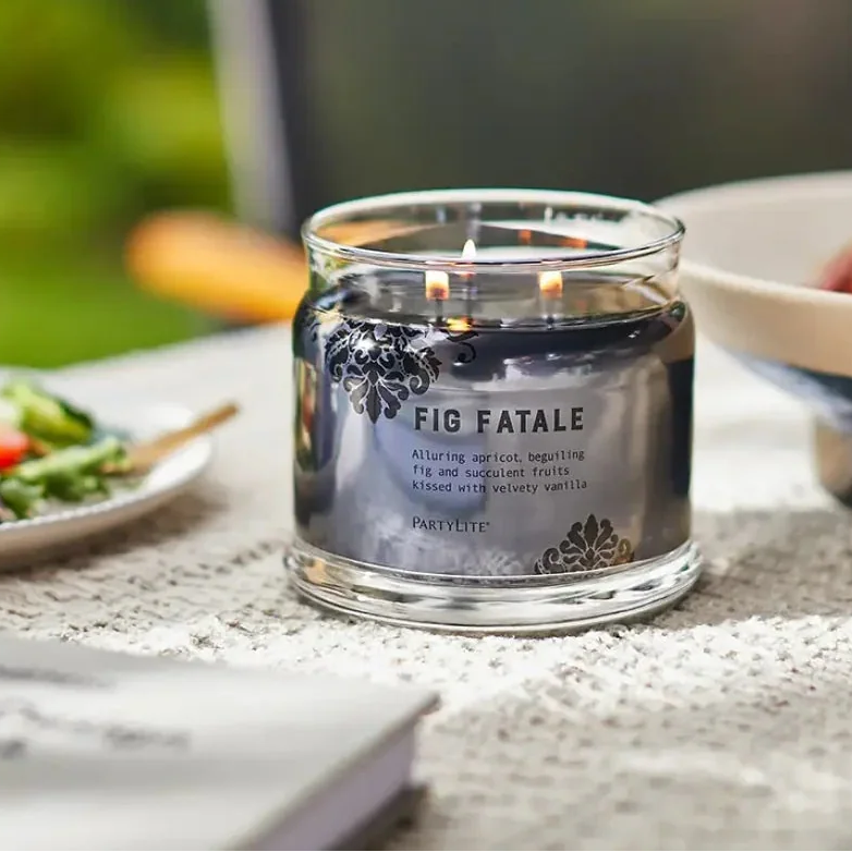 Lit dark brown 3-wick candle on table next to bowl of fresh figs and plated salad.