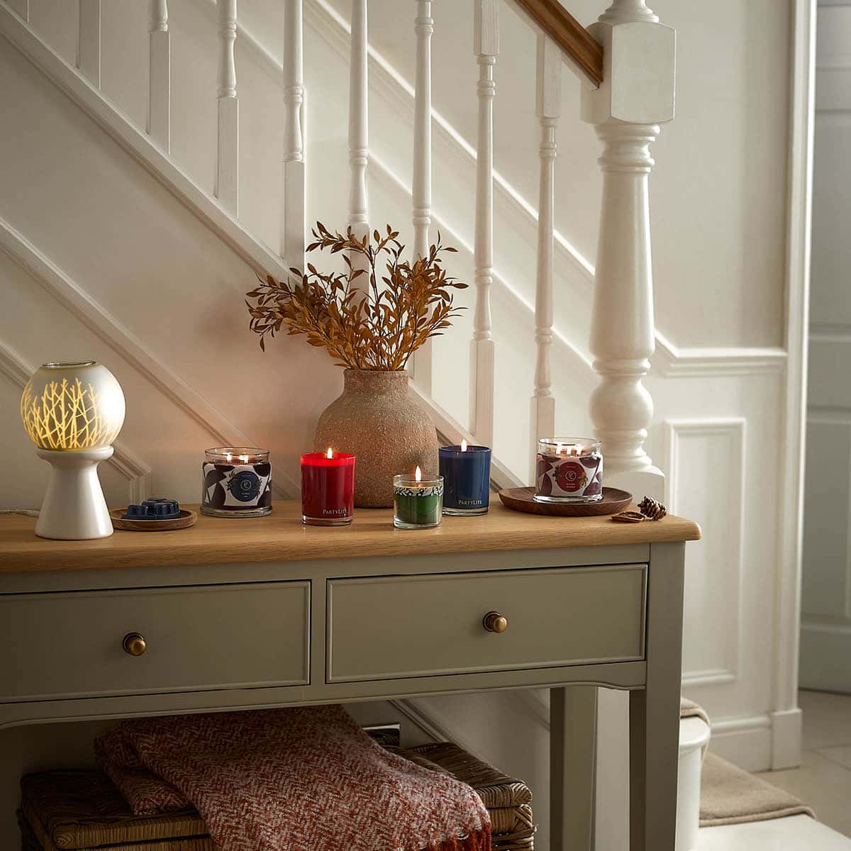 Wild Woods & Foliage Mini Jar Candle shown lit with other PartyLite Escential Jars, a 3-wick and a scent glow warmer on a cabinet with stairs in the background.