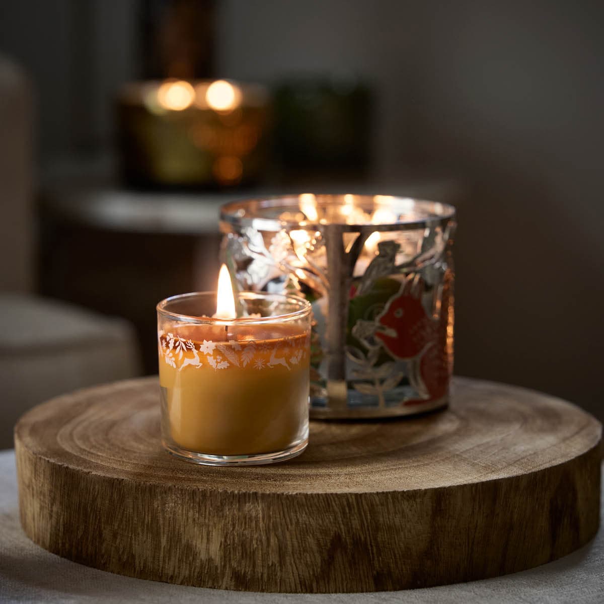 Whiskey & Oak Mini Jar Candle lit shown with the Squirrel Jar Holder on a round wooden surface with a blurred background.