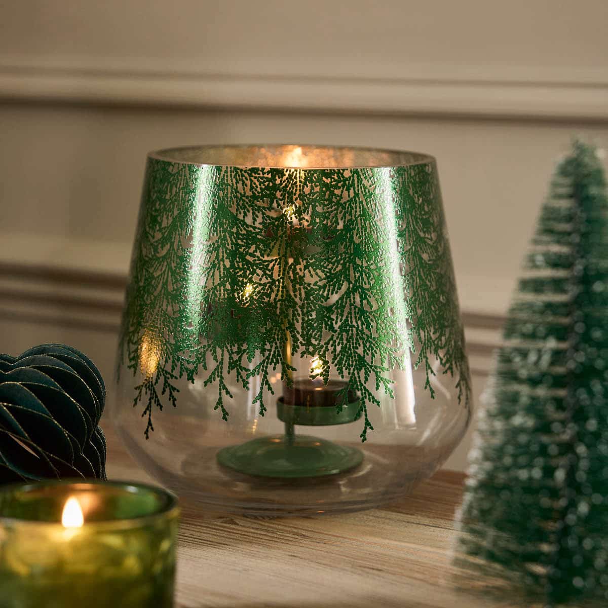 A clear glass hurricane with a metallic finish, adorned with a lace-like pattern of evergreen branches. It includes a metal tealight holder shown with a lit tealights on a wooden surface.