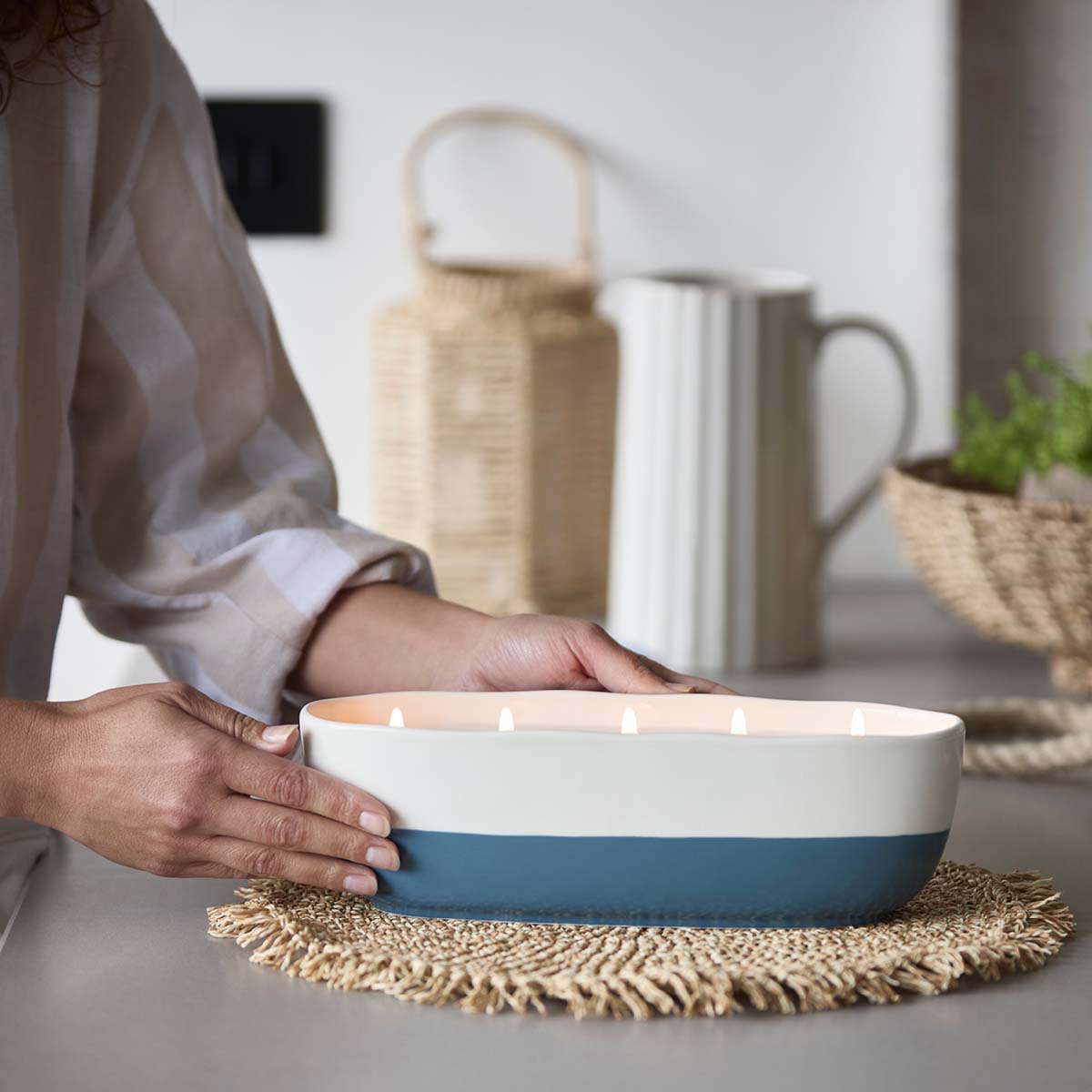 Capri Limoncello Specialty Jar Candle, with 5 wicks with a white top and blue bottom ceramic holder, with a ladies hands either side on a woven table mat.