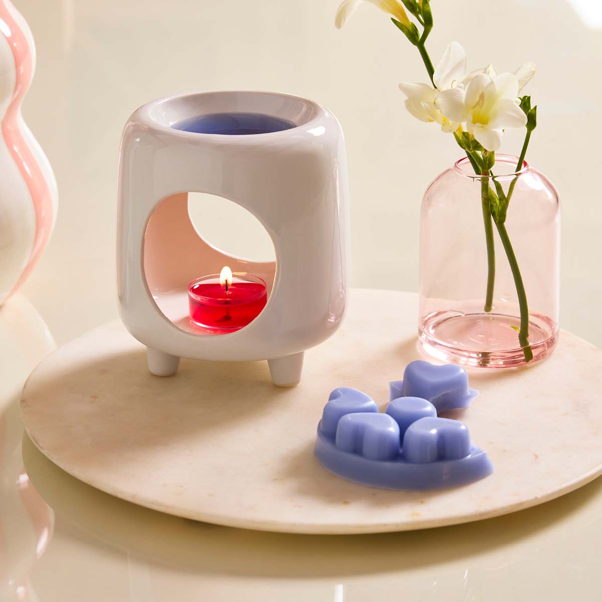 Mini Chiminea Fragrance Warmer shown with heart shaped wax melts on a light colored tray with a clear rose colored vase with white flowers.