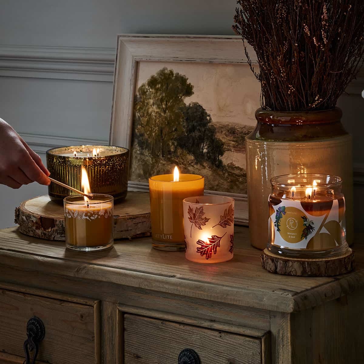 Whiskey & Oak Mini Jar Candle being lit by a long matchstick shown with other PartyLite jar candles also lit on a wooden cabinet and a picture frame with a tree in the background.