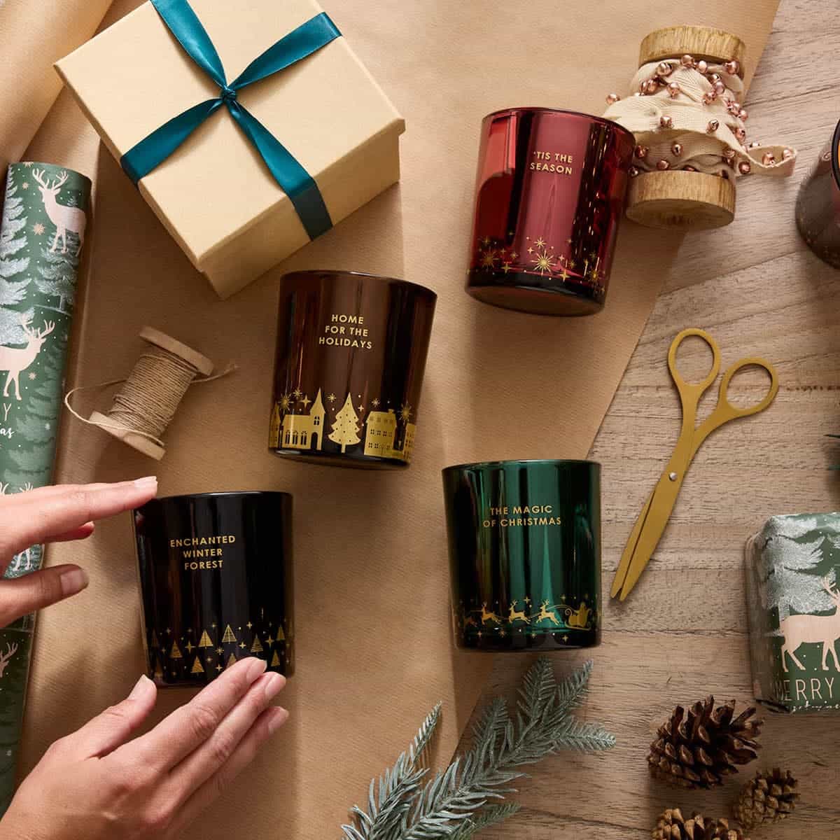 Assortment of Holiday Themed colorful jars in various fragrances, shown on a wooden table with brown wrapping paper and scissors.