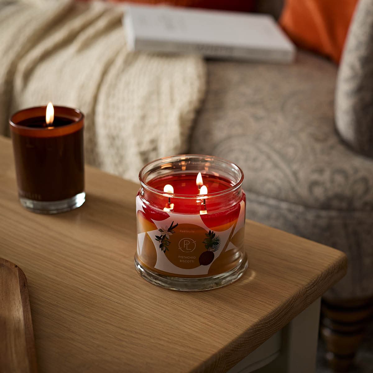Pistachio Biscotti 3-wick Jar Candle lit on a wooden table with an escential jar also lit and a chair in the background.