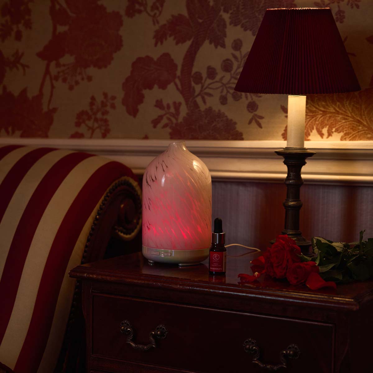 Passionate Fragrance Oil from the Mood Collection shown next to a lamp and Red Roses on a chest of drawers and a fragrance diffuser with a stripy red and cream chair cushion next to it.
