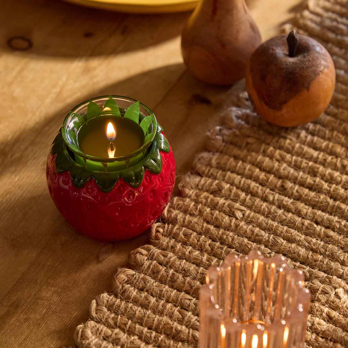 Strawberry Tealight/Votive Holder shown with a lit candles with a woven table mat next to it on a wooden surface.