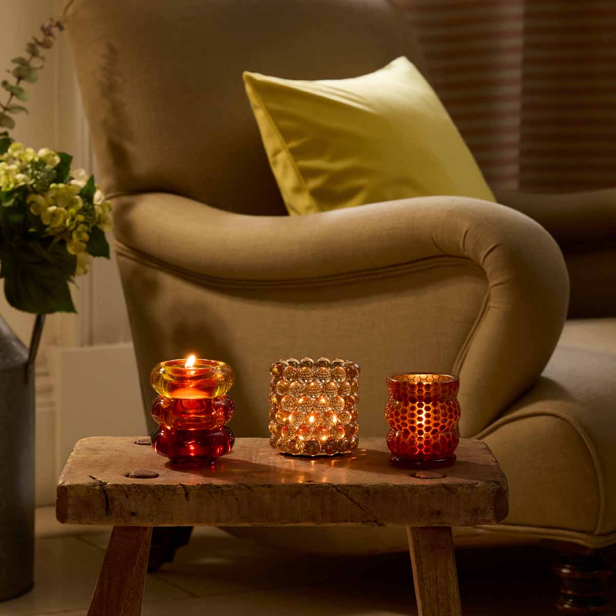 Glass Bead Tealight Holder shown lit between other Tealight holders on a wooden table with an armchair next to it.