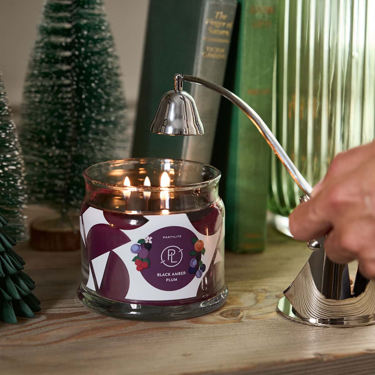 Black Amber Plum 3-Wick Jar Candle lit with a hand holding a chrome snuffer hoovering above on a wooden surface.