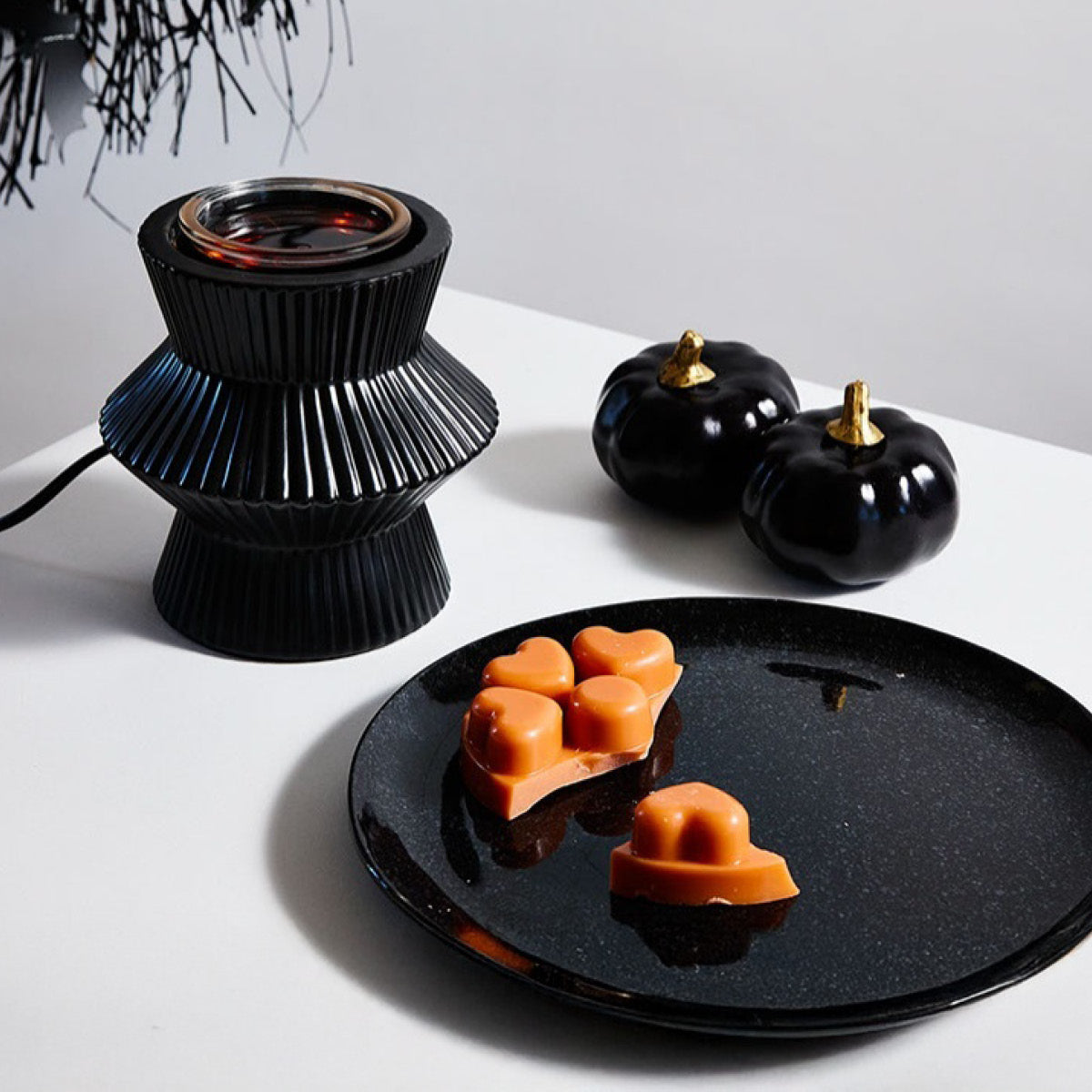 Spiced Pumpkin heart shaped wax melts on a black plate with a black scent glow warmer in next to it with black decorate pumpkins on a white background