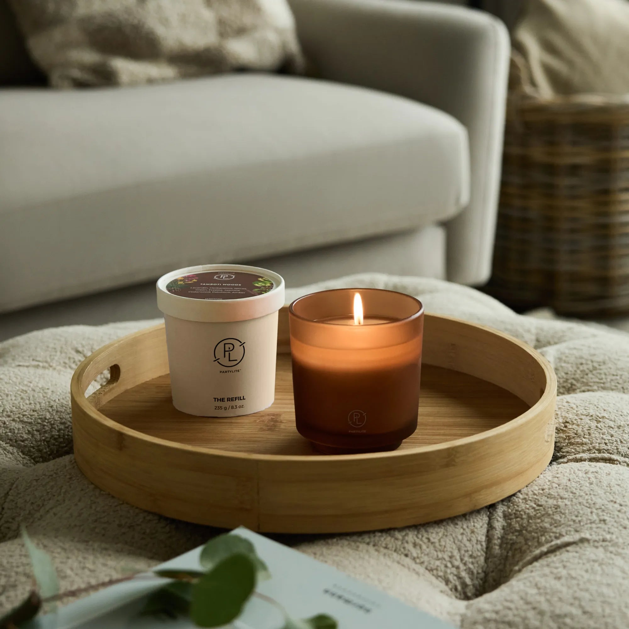Tamboti Woods Refillable Jar Candle on a wooden tray with a refill shown next to it on a cushioned surface with a sofa in the background