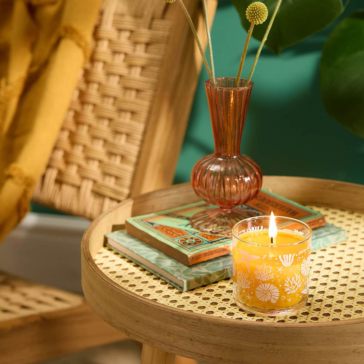 Sun Orchid Pineapple Mini Jar Candle on a small wooden table with books and clear brown vase of flowers next to it with an outline of woven chair with a green background