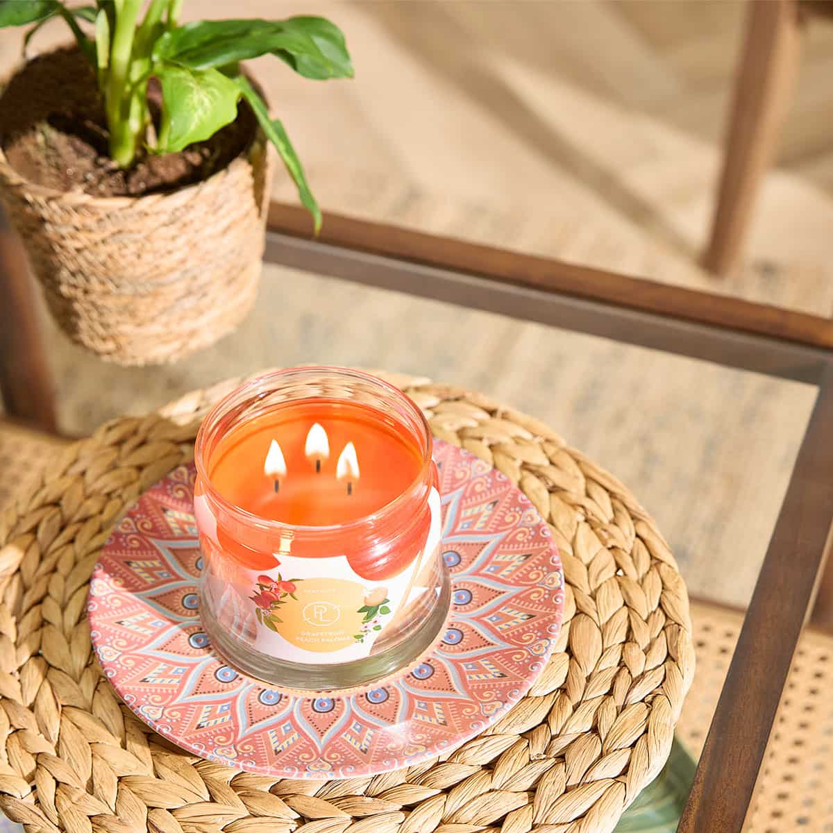 Grapefruit Peach Paloma 3-Wick Jar Candle shown on a decorative plate on a woven round mat on a glass table table top with a plant in the background