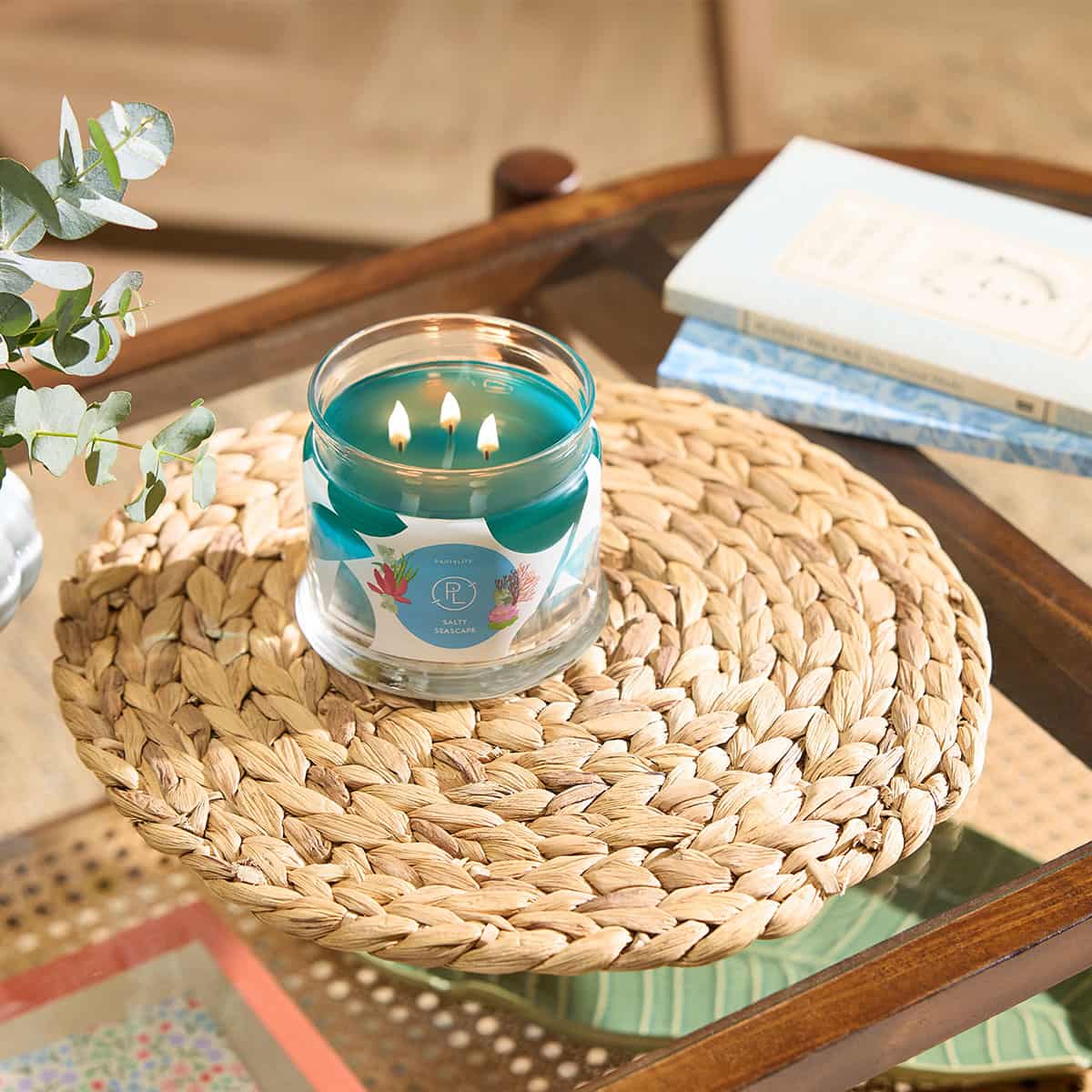 Salty Seascape 3-wick Jar Candle lit on a woven table mat on a glass table top with books lying on the glass top with a beige background