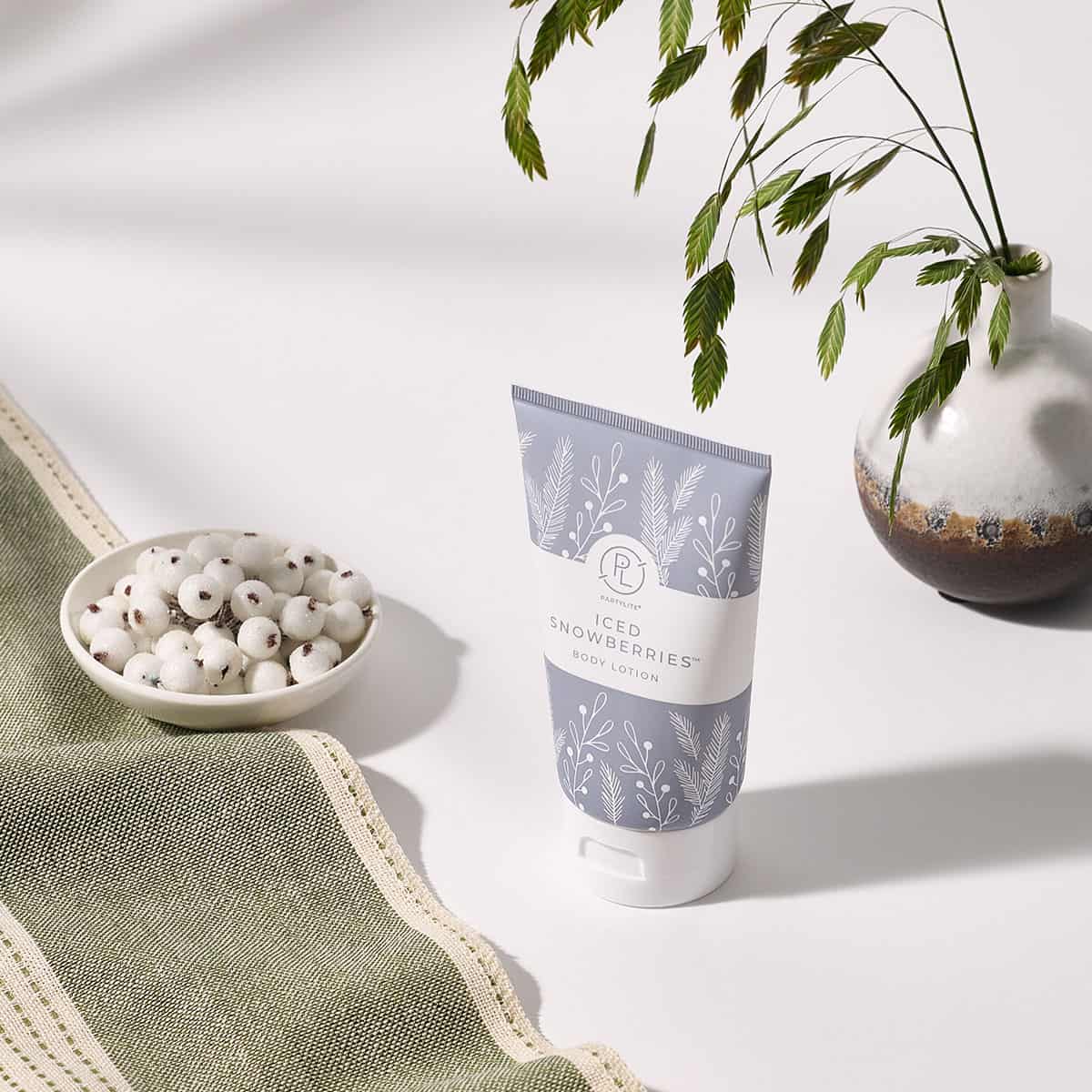 Iced Snowberries Body Lotion with a small bowl of white berries and a ceramic vase with leaves in the background on a white surface