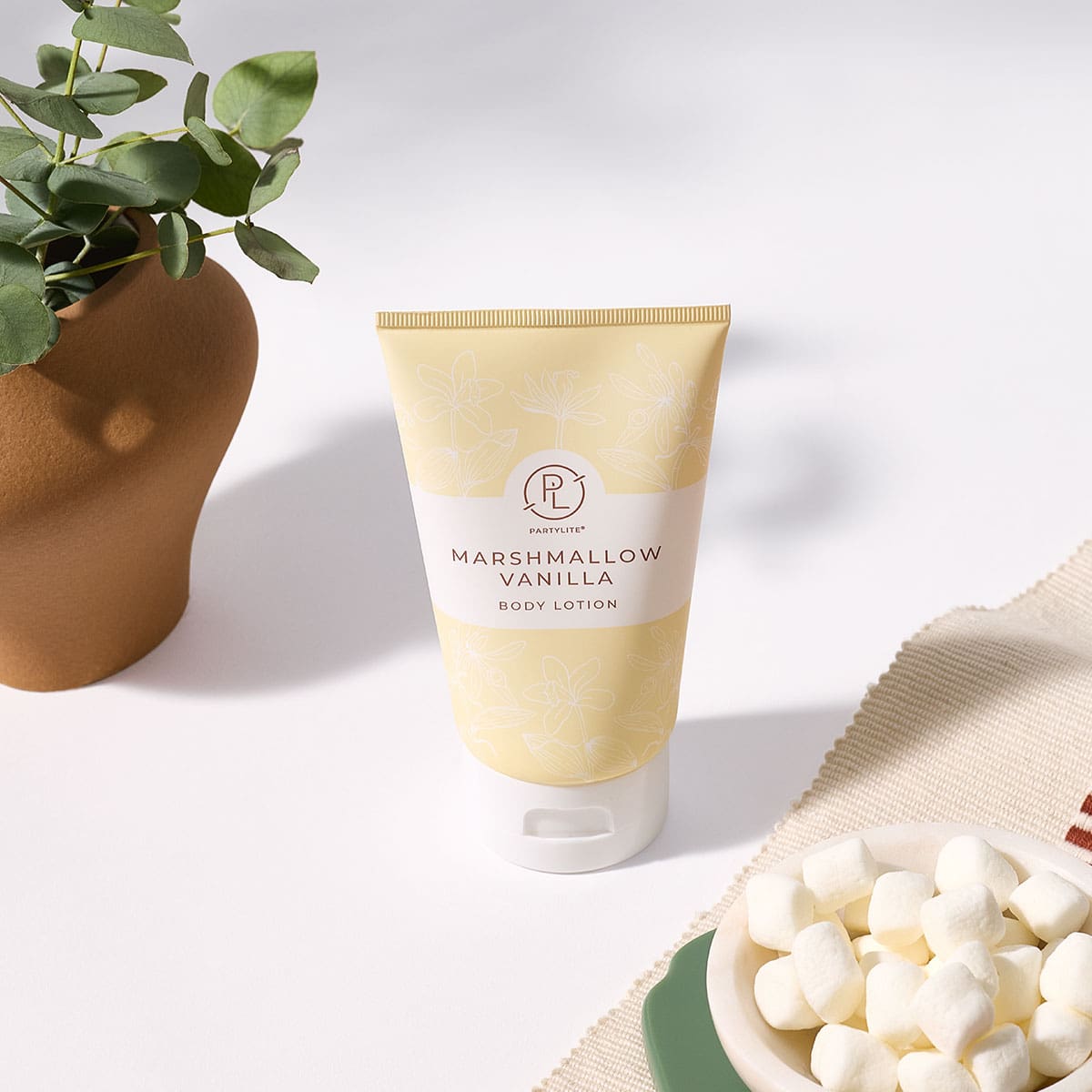 Marshmallow Vanilla Body Lotion on a white surface next to a plant and a small bowl of Marshmallows