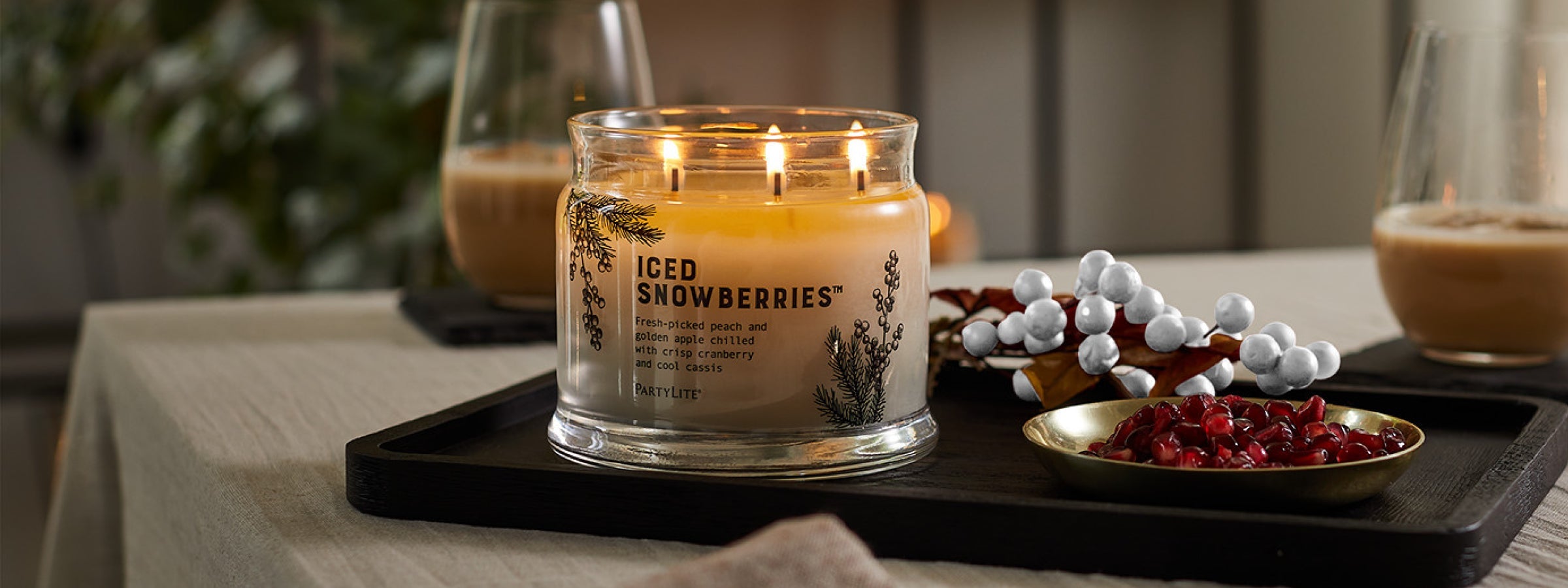 Lit off-white 3-wick candle on a tray next to red and white berries with two beverages in the background.