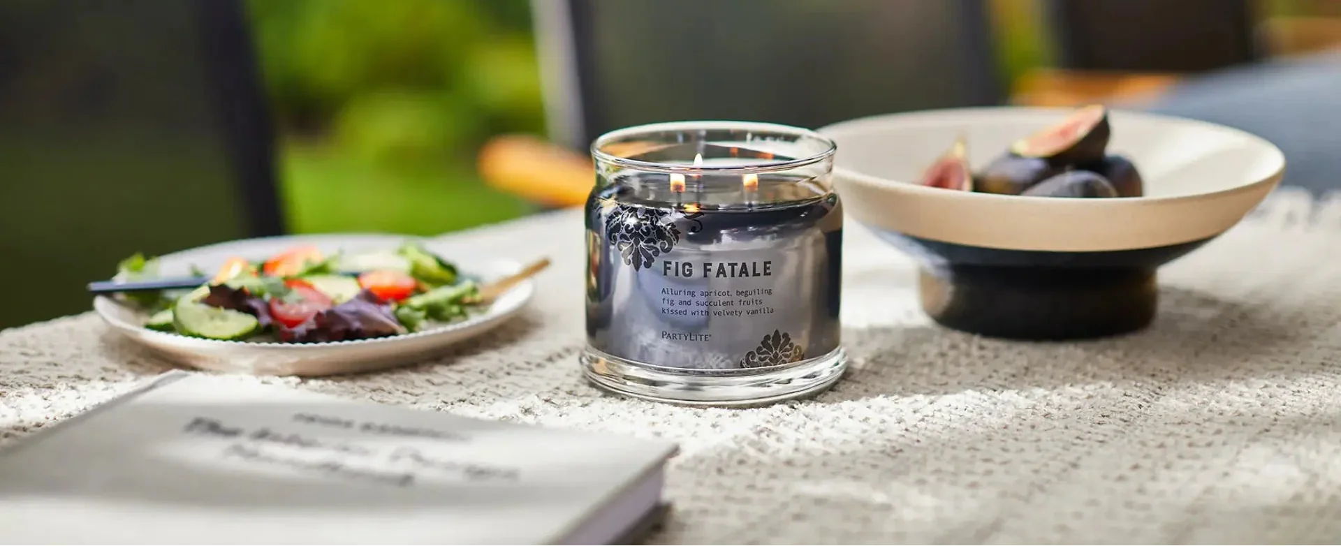 Lit dark brown 3-wick candle on table next to bowl of fresh figs and plated salad.