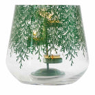 A clear glass hurricane with a metallic finish, adorned with a lace-like pattern of evergreen branches. It includes a metal tealight holder shown with a lit tealights.