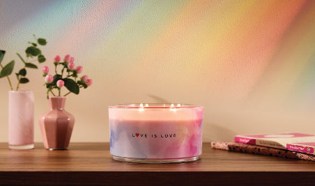 Candle with 'Love is Love' text on a wooden surface with vases and flowers in the background