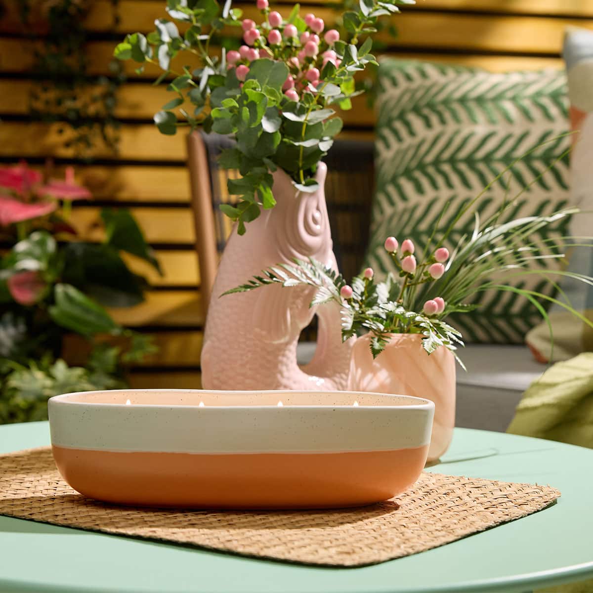 Peach Honey Citronella 4-wick two tone Jar Candle in a garden surroundings on a woven table mat on a small green table with vases of flowers and potted plant in the background with an outdoor green and cream cushion on a garden sofa