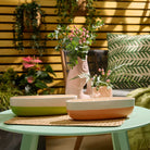 Peach Honey Citronella 4-wick two tone Jar candle shown with Wild Lemongrass Citronella 4-wick tone tone Jar Candle in garden setting, sitting on a woven table mat on a small green table with a vase of flowers with hanging plants in the background